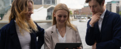 Drei Studierende in Business-Outfits unterhalten sich und schauen auf ein Tablet.